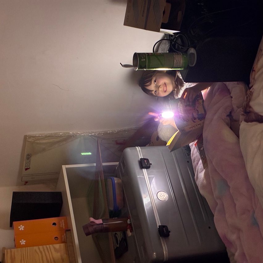 Ein Kind hält eine Taschenlampe in einem Zimmer. Ein Koffer steht auf dem Boden neben einem Regal.