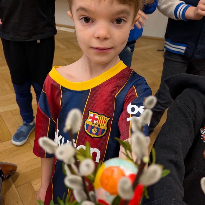 Ein Junge in einem FC Barcelona Trikot, das ein verziert Ei hält, steht unter anderen in einem Raum mit Holzboden.