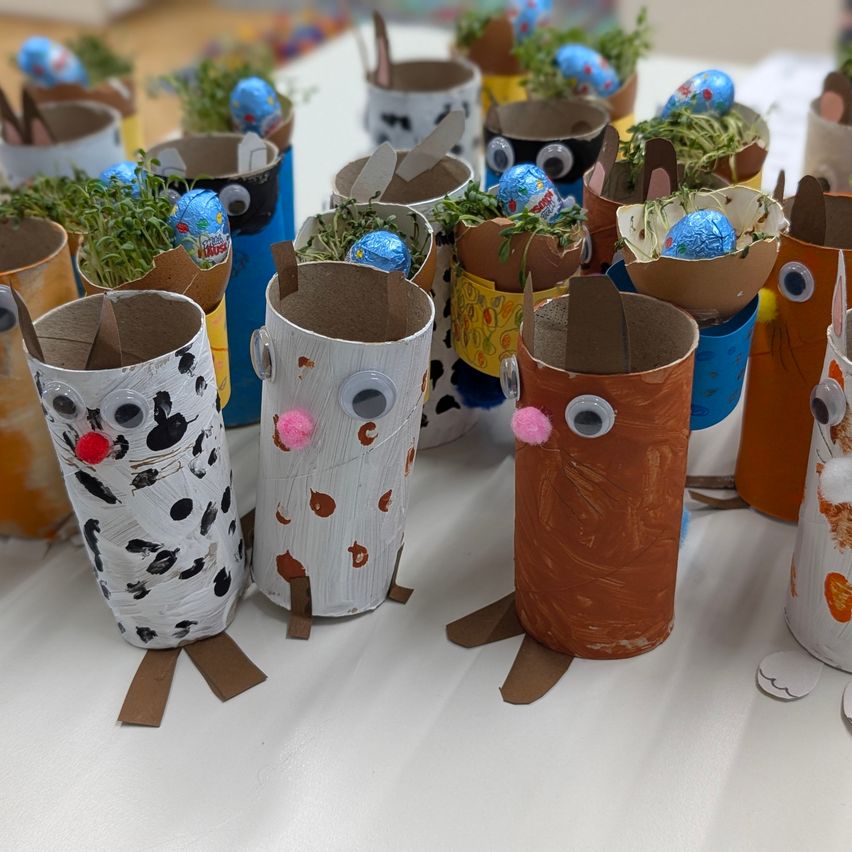 Ein Tisch mit zahlreichen dekorierten Pappröhren, jede mit einem Osterei und grünen Sprossen darin, in einer Reihe angeordnet.