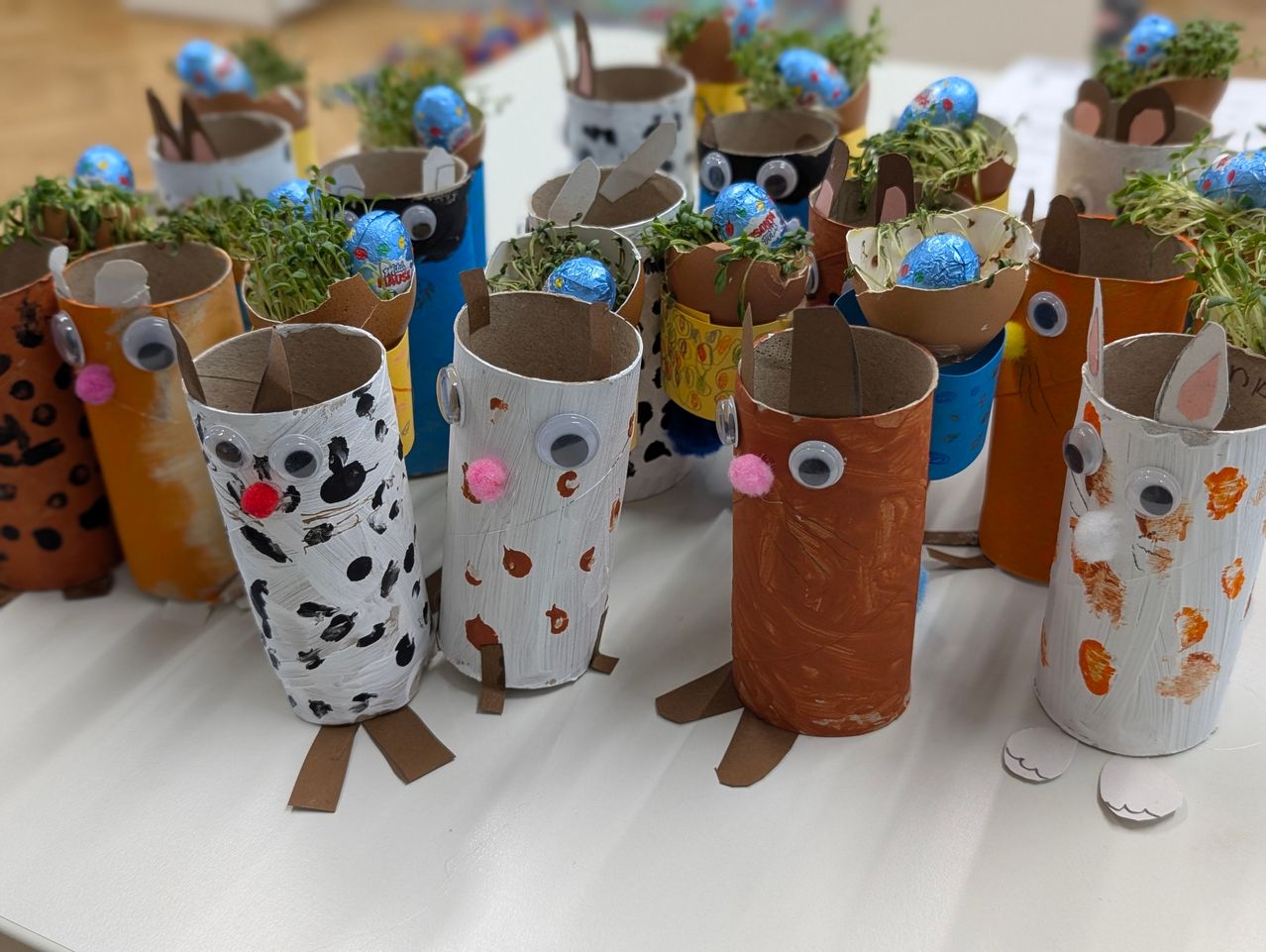 Ein Tisch mit zahlreichen dekorierten Pappröhren, jede mit einem Osterei und grünen Sprossen darin, in einer Reihe angeordnet.