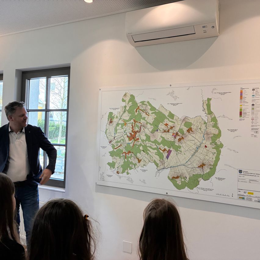 Ein Mann zeigt Kindern eine Karte. Die Karte zeigt verschiedene farbige Bereiche, die möglicherweise verschiedene Gebiete oder Zonen darstellen. Die Umgebung ist innen mit einem Fenster und einer Klimaanlage an der Wand.