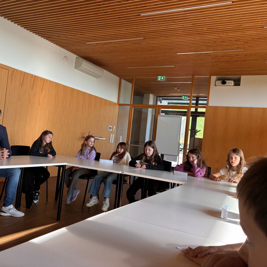 Eine Gruppe junger Mädchen sitzt an einem Tisch in einem Konferenzraum. Ein Mann steht vorne, möglicherweise in einer Diskussion. Der Raum ist gut beleuchtet und hat Holzwände und -decken.