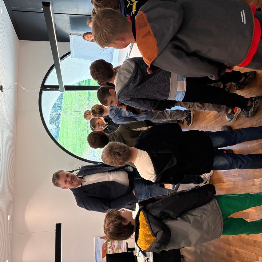 Eine Gruppe von Kindern und ein erwachsener Mann stehen in einem Raum mit Holzboden. Sie schauen alle nach unten, wahrscheinlich zu einer Ausstellung. Im Hintergrund befindet sich ein großes Fenster.