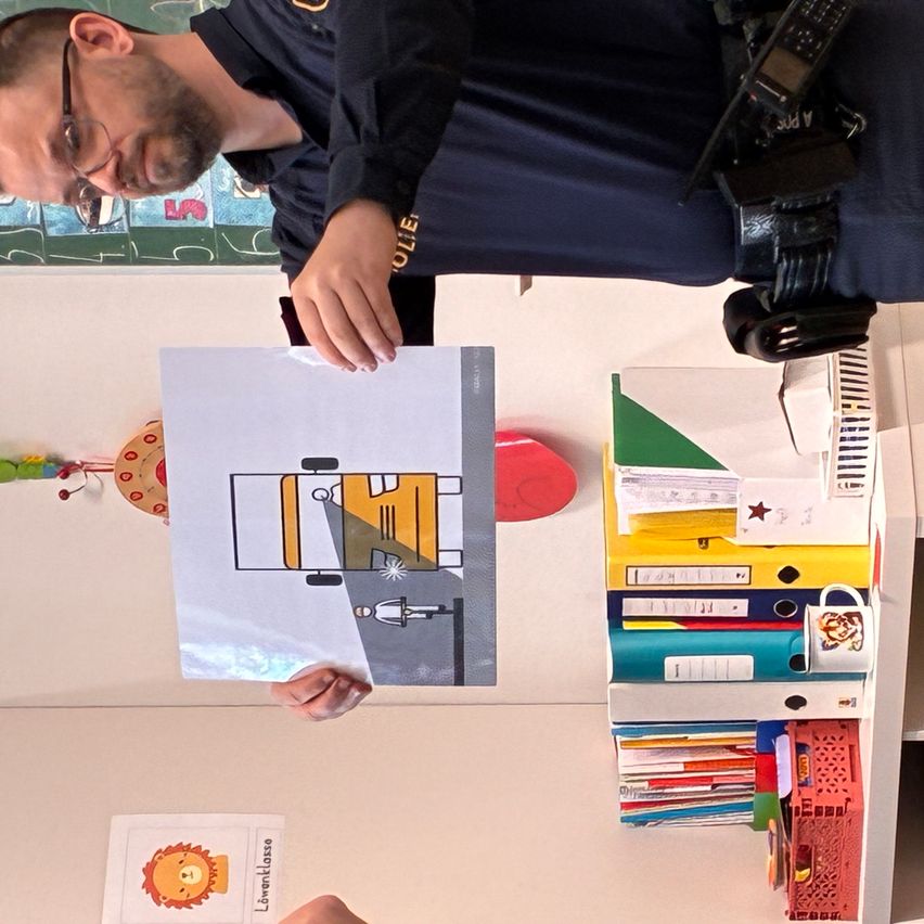 Ein Mann mit Brille und einem marineblauen Hemd hält ein Bild eines Fahrzeugs. Vor ihm steht ein Schreibtisch mit mehreren Ordnern, einem Becher und einer Stiftbox.