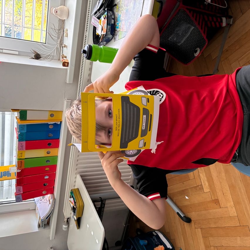 Ein Junge in einem roten Shirt hält einen gelben Papierbus hoch. Er sitzt auf einem Stuhl in einem Klassenzimmer mit Holzböden und Regalen mit Ordnern.