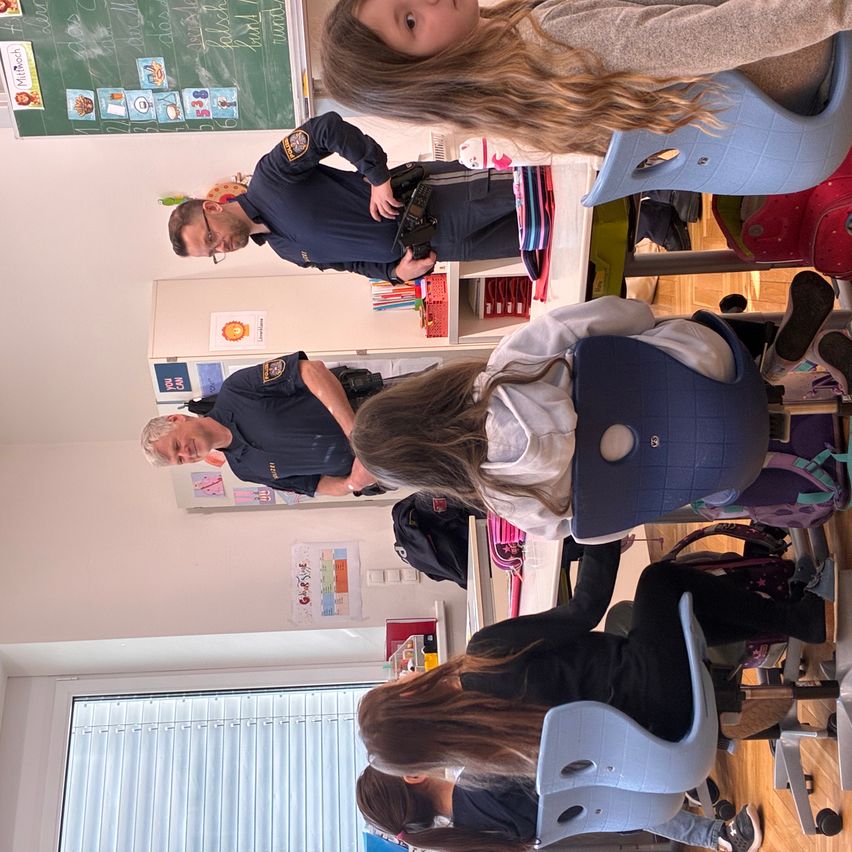 Ein Polizist steht in einem Klassenzimmer mit Schülern. Er hält eine Kamera. Es gibt Tische und Stühle im Raum.