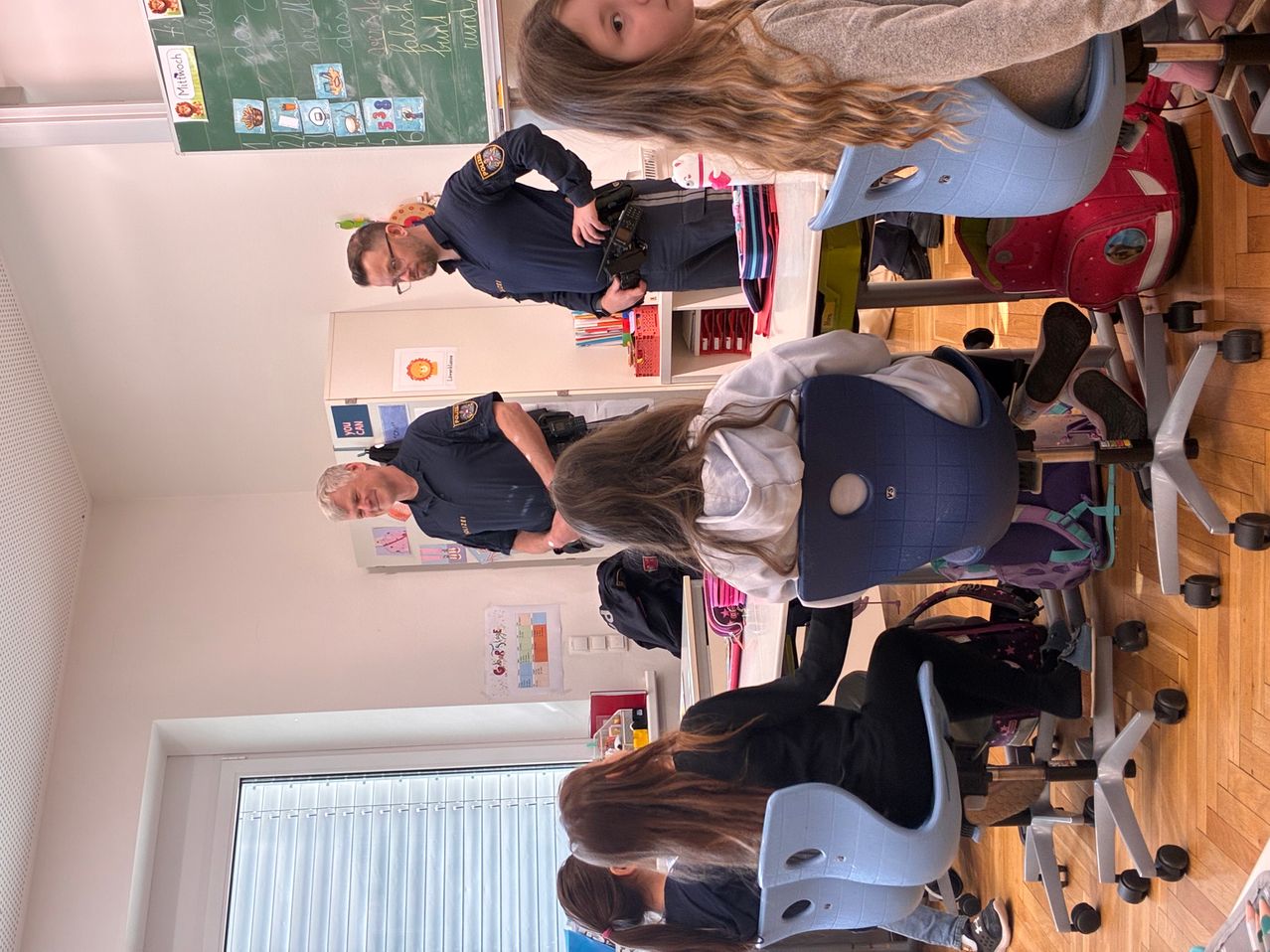 Ein Polizist steht in einem Klassenzimmer mit Schülern. Er hält eine Kamera. Es gibt Tische und Stühle im Raum.