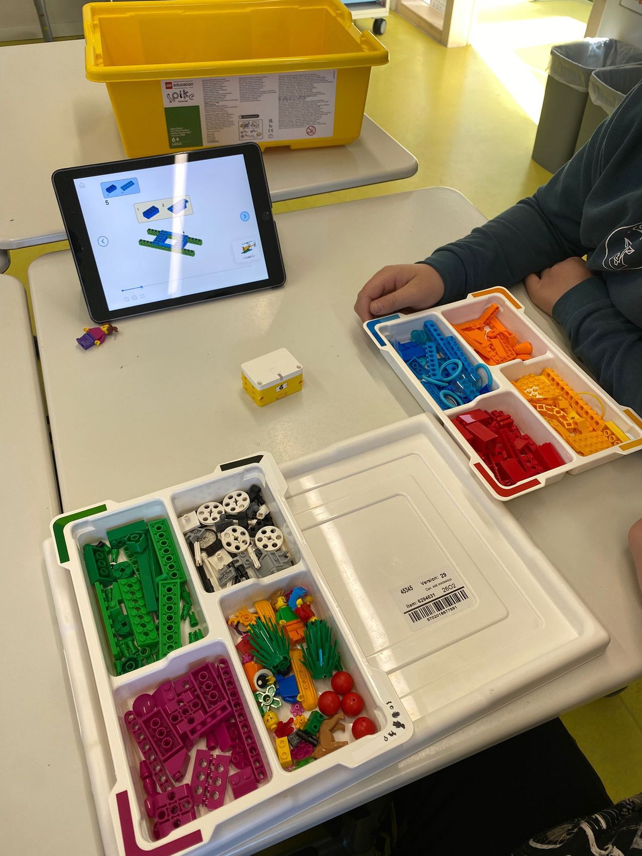 Eine Person sitzt an einem Tisch und benutzt ein Tablet, um sie beim Bauen mit bunten Legosteinen zu leiten. Eine Lego-Minifigur und eine gelbe Box sind in der Nähe.