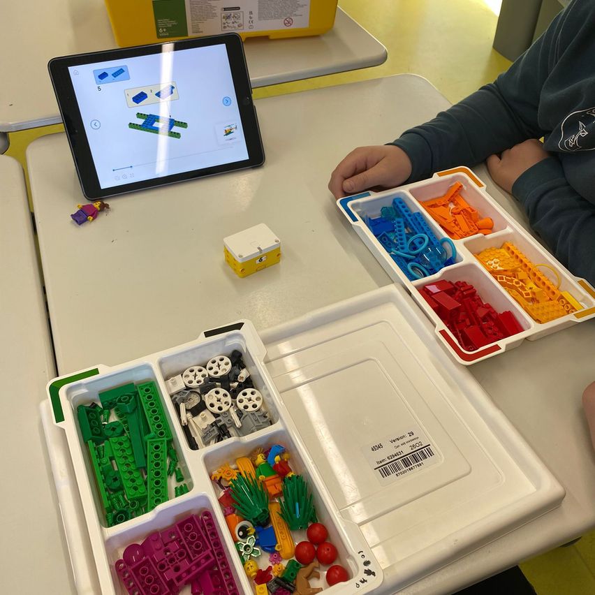 Eine Person sitzt an einem Tisch und benutzt ein Tablet, um sie beim Bauen mit bunten Legosteinen zu leiten. Eine Lego-Minifigur und eine gelbe Box sind in der Nähe.