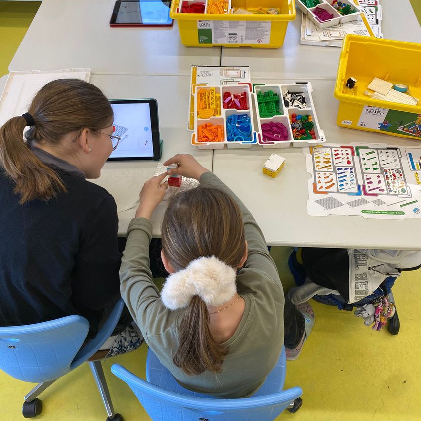 Zwei Mädchen sitzen an einem Tisch und arbeiten gemeinsam an einem Projekt mit bunten Bausteinen und einem Tablet als Anleitung.