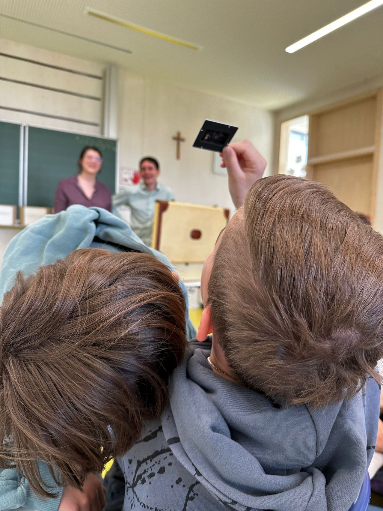 Zwei Jungen in einem Klassenzimmer machen ein Foto mit einer Kamera. Eine Lehrerin und ein Mann sind im Hintergrund zu sehen.