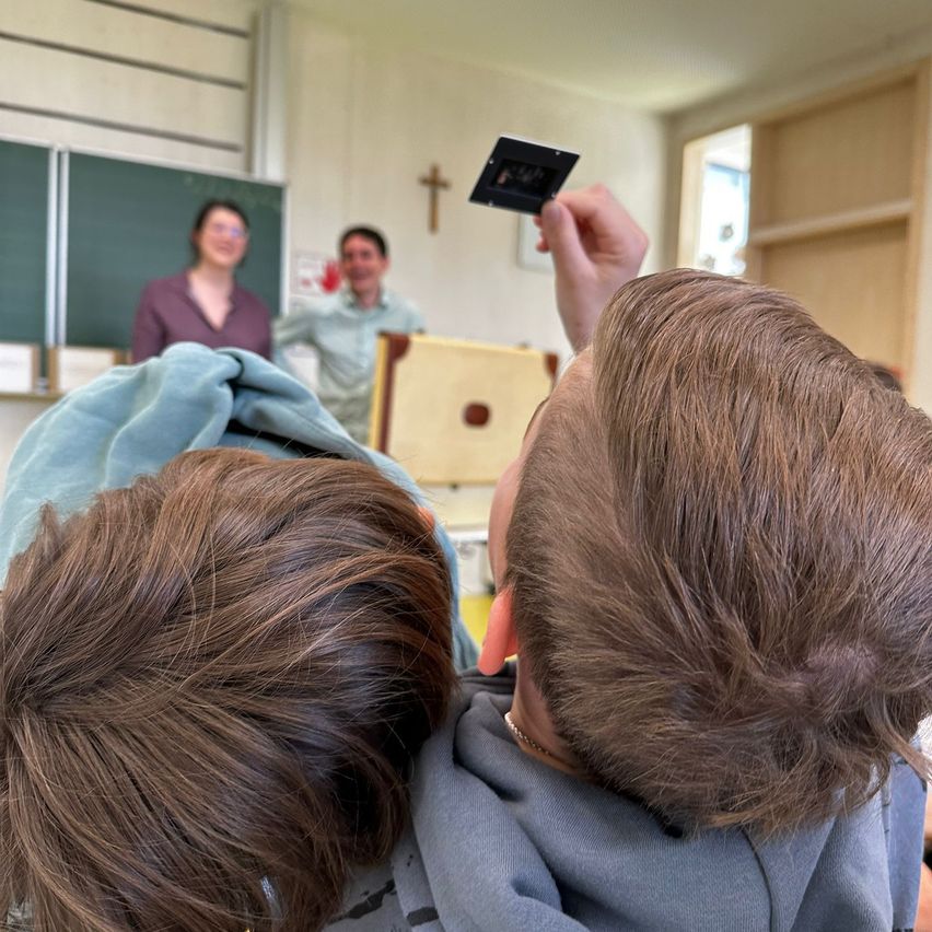 Zwei Jungen in einem Klassenzimmer machen ein Foto mit einer Kamera. Eine Lehrerin und ein Mann sind im Hintergrund zu sehen.
