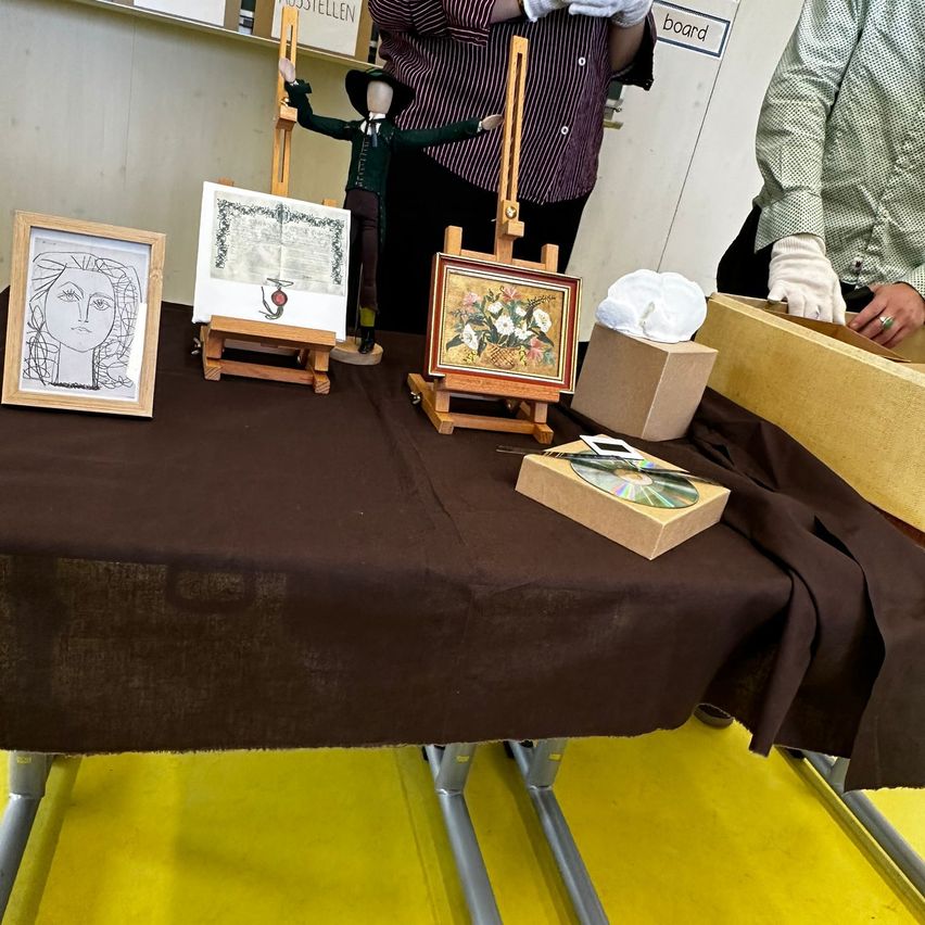 Ein mit einem braunen Tuch bedeckter Tisch zeigt mehrere Kunstwerke. Ein Frauenportrait, ein gerahmtes Gemälde, ein Zertifikat und ein CD-Karton liegen auf dem Tisch. Eine Person steht hinter dem Tisch.