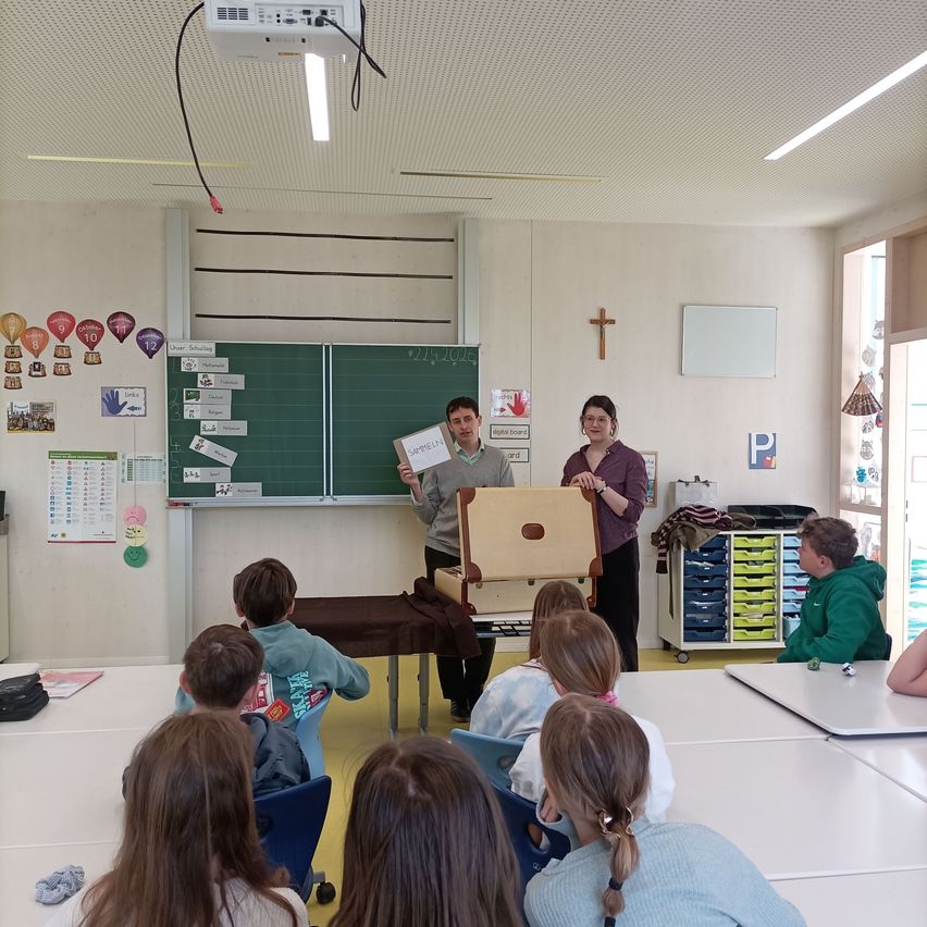 Ein Klassenzimmer mit einem Lehrer und einem Schüler, die eine Aktentasche präsentieren. Viele Kinder sitzen auf Stühlen und schauen zu. Eine Tafel, Tische und ein Projektor befinden sich im Raum.