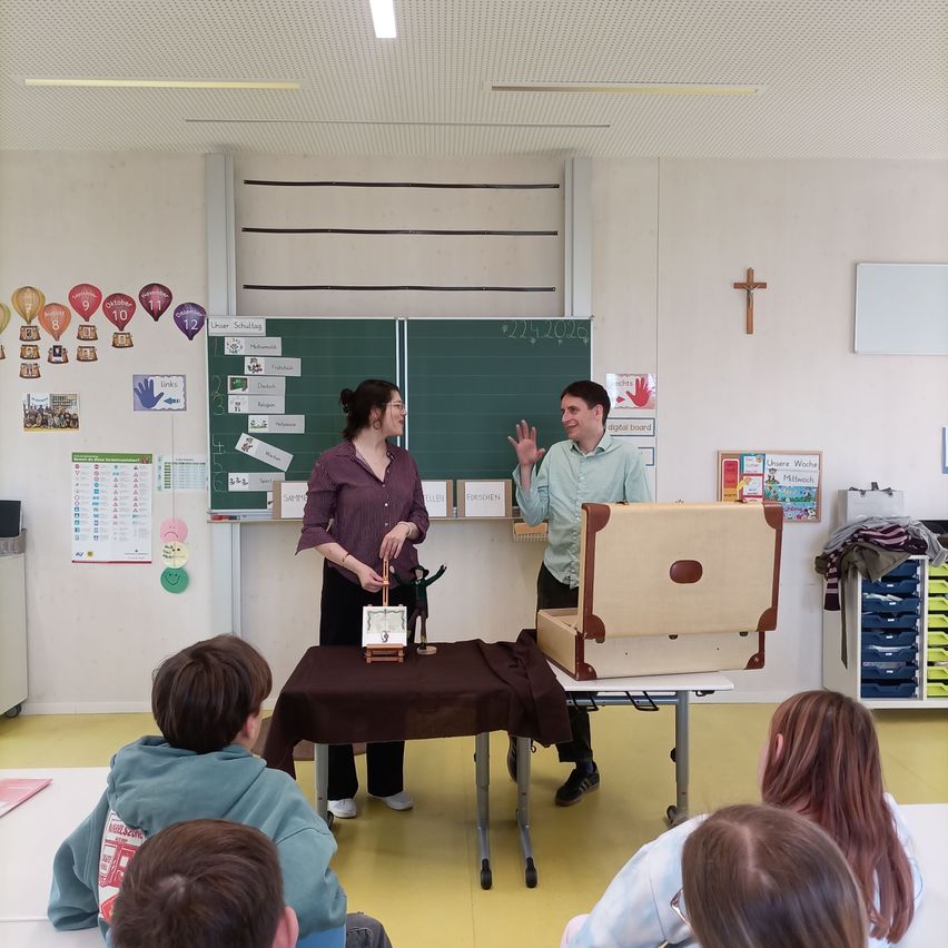 Ein Klassenzimmer mit einer Tafel zeigt verschiedene Dekorationen. Ein Mann und eine Frau stehen vor einem Tisch mit einem offenen Koffer und einem Tuch, vielleicht führen sie einen Zaubertrick für eine Gruppe von Kindern vor.
