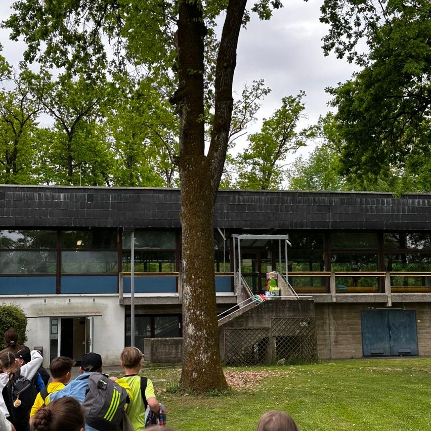 Eine Gruppe von Kindern mit Rucksäcken und Mützen versammelt sich vor einem modernen Gebäude mit Glasfenstern. Sie scheinen darauf zu warten, dass sie eintreten können.