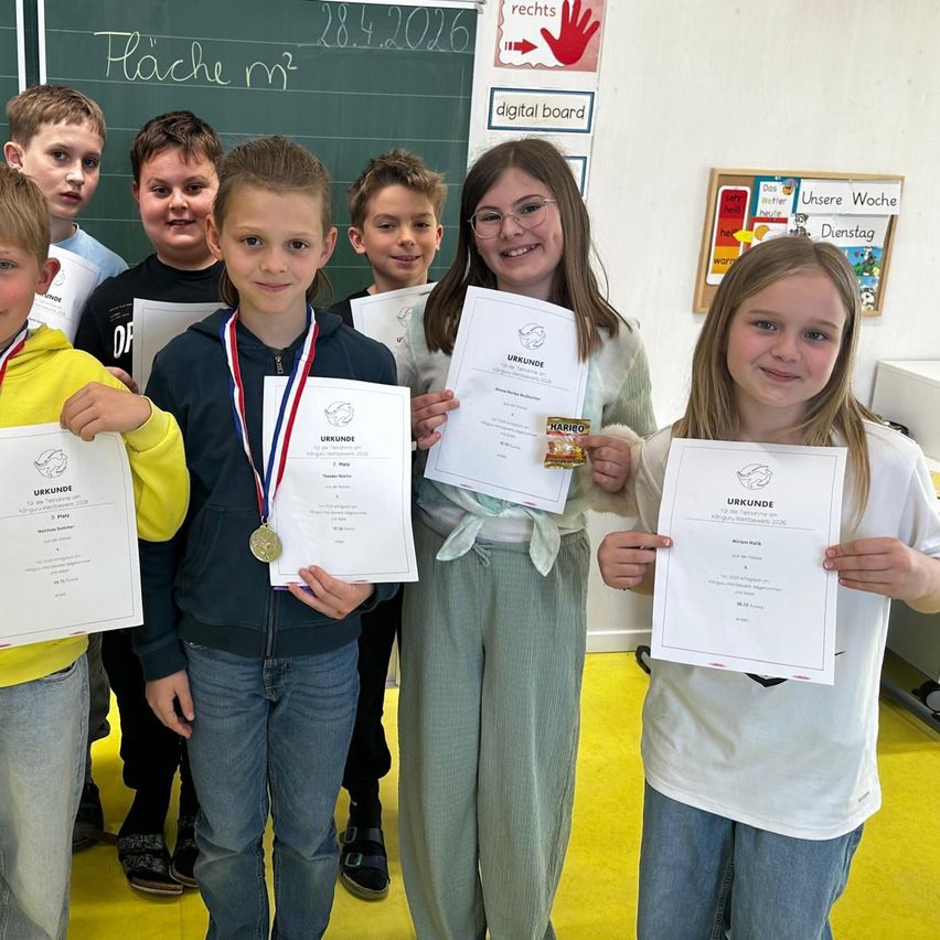 Gruppe von Kindern in einem Klassenzimmer mit Urkunden und Medaillen. Ein Schultafel hinter ihnen hat Text und eine Hinweistafel für ein digitales Board. Sie lächeln.
