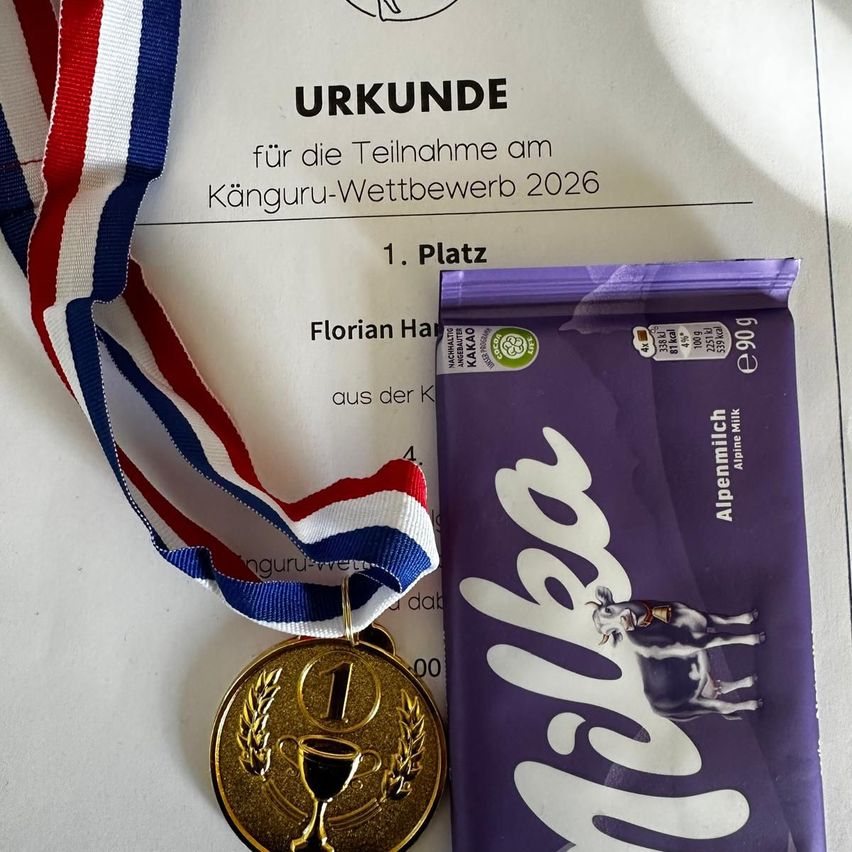 Eine goldene Medaille mit '1' und einem Pokalsymbol darauf wird neben einer lila Alpenmilch-Schokoladentafel gezeigt. Die Medaille ist an einem Band mit roten, weißen und blauen Streifen befestigt.