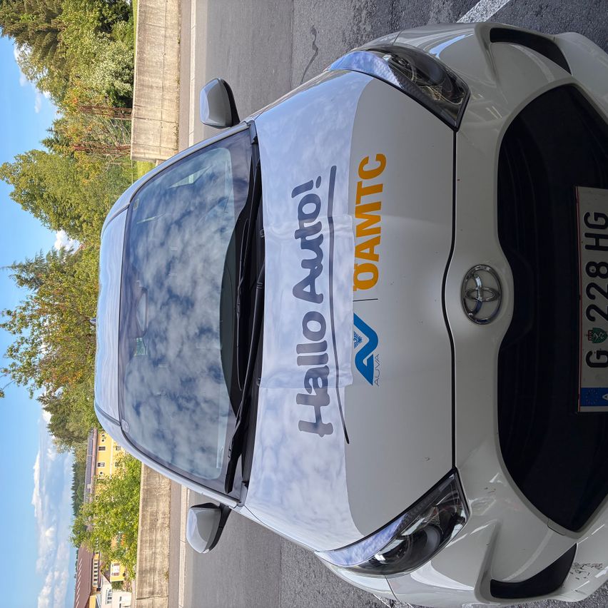 Ein weißer Wagen mit den Worten 'Hallo Auto!' und 'OAMTC' auf der Motorhaube, geparkt am Straßenrand.