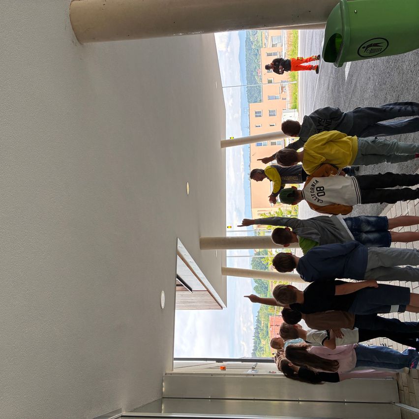 Eine Gruppe von Kindern und einer Person in orangefarbener Hose schaut von einem Balkon hinunter. Ein grüner Mülleimer ist in der Nähe. Die Kinder tragen Kleidung in verschiedenen Farben und Größen.