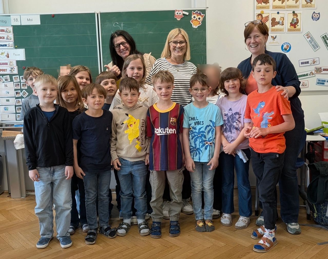 Eine Gruppe von Kindern und drei Frauen steht in einem Klassenzimmer und lächelt. Die Kinder tragen Freizeitkleidung, und die Frauen tragen Brillen und Freizeitkleidung. Hinter ihnen steht eine Tafel mit einigen Zeichnungen.