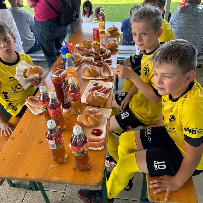 Vier Jungen in gelben Trikots sitzen an einem Tisch und essen Hot Dogs. Verschiedene Gewürze und Getränke stehen auf dem Tisch. Hinter ihnen sitzen Menschen auf Bänken, einige mit Rucksäcken.