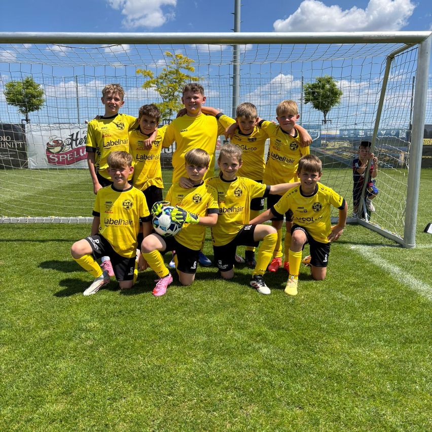 Eine Jugendfußballmannschaft in gelben Trikots posiert für ein Foto auf einem Rasen. Sie knien und sitzen, halten einen Fußball. Dahinter steht ein Tornetz und ein Junge steht vor einem Banner.