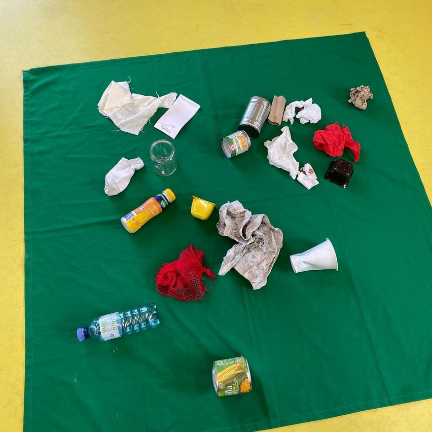 Verschiedene Abfallmaterialien wie Plastikflaschen, Dosen, Papiertücher und Taschentücher sind auf einem grünen Tuch verteilt. Es gibt zwei Plastikflaschen, von denen eine mit 'Villa' und eine Dose mit 'Olla' beschriftet ist.