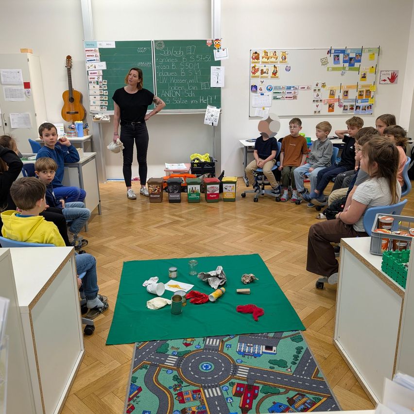Eine Frau steht vor einem Klassenzimmer und erklärt etwas, während Kinder auf Stühlen sitzen und aufmerksam zuhören. Ein Tafel und eine Tafel sind im Hintergrund sichtbar.