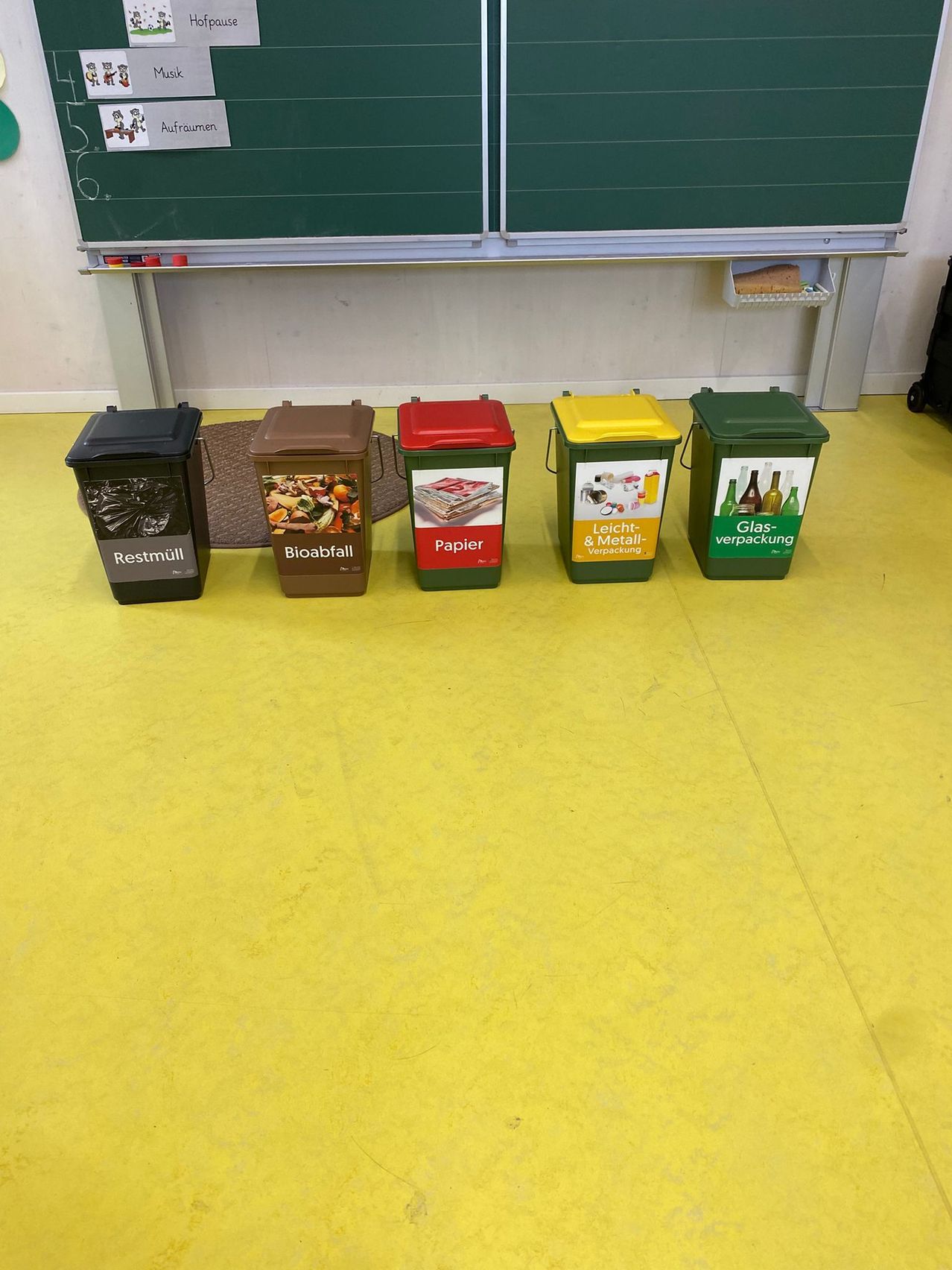 Fünf Recyclingbehälter stehen auf einem gelben Boden. Von links sind die Behälter für Restmüll, Bioabfall, Papier, Leichtmetall und Glas vorgesehen.