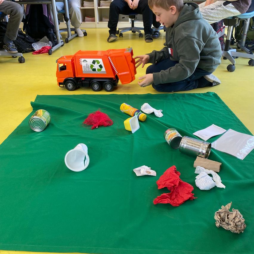 Ein junger Junge spielt mit einem Spielzeug-Recycling-Lkw auf einer grünen Matte. Verschiedene recycelbare und Abfallgegenstände sind um ihn herum verstreut.