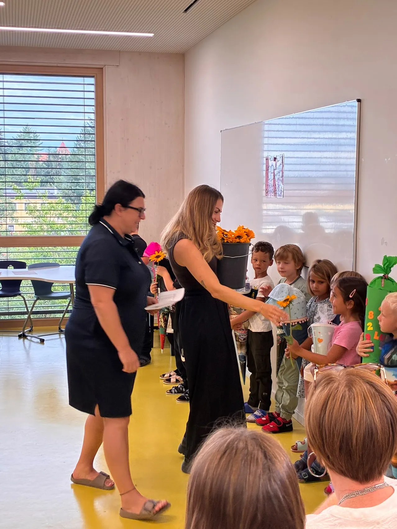 Eine Frau in einem schwarzen Kleid verteilt Blumen an Kinder in einem Klassenzimmer.