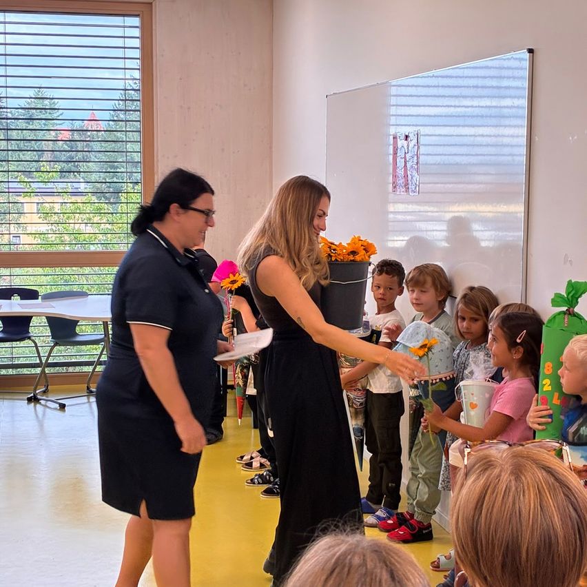 Eine Frau in einem schwarzen Kleid verteilt Blumen an Kinder in einem Klassenzimmer.