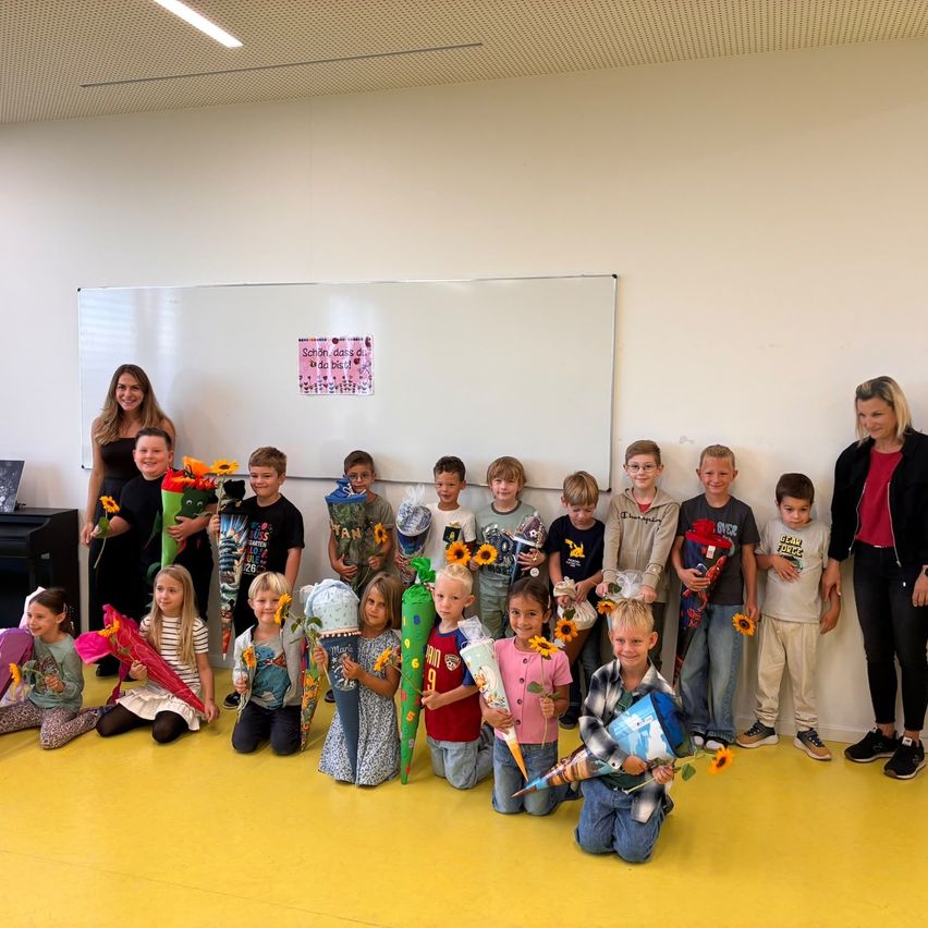 Eine Gruppe von Kindern und zwei Frauen posiert für ein Foto im Klassenzimmer, hält Blumen und trägt Freizeitkleidung.