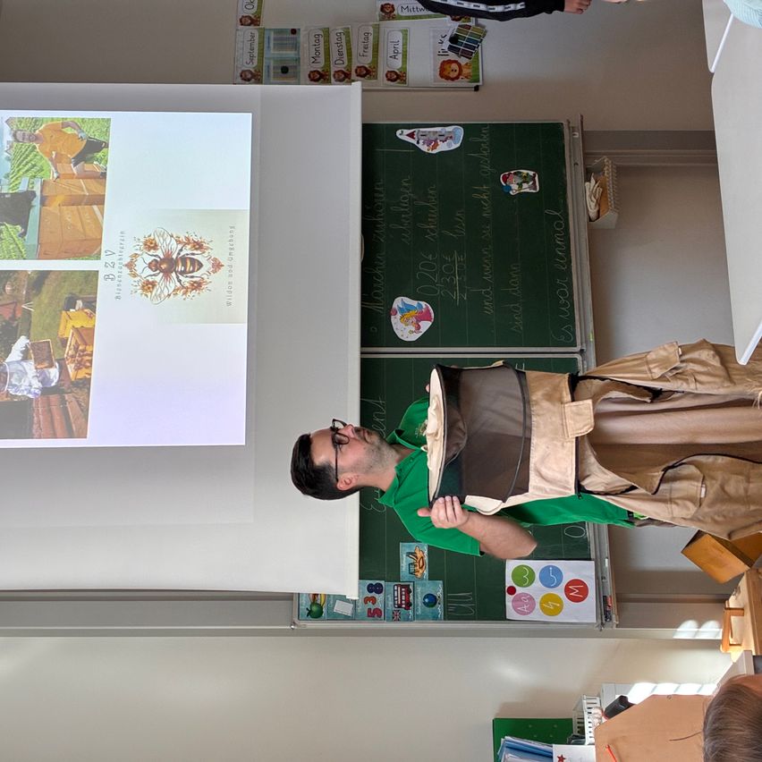 Ein Mann in einem grünen Shirt und einer Imkerausrüstung steht in einem Klassenzimmer mit einer Projektorleinwand, die Bienenbilder zeigt.