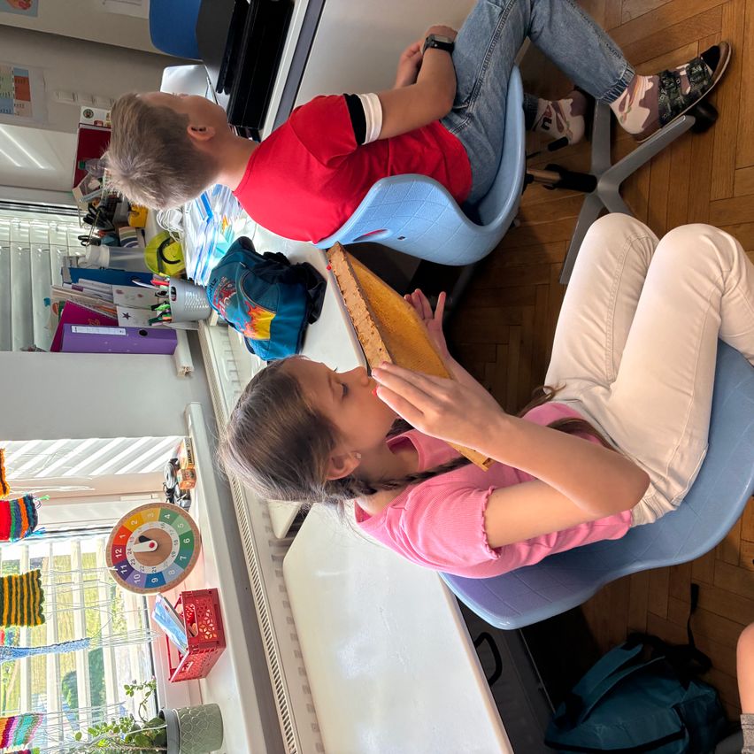 Ein Junge und ein Mädchen sitzen in Stühlen, wahrscheinlich in einem Klassenzimmer. Das Mädchen isst ein Stück Kuchen, während der Junge zusieht. Hinter ihnen steht ein Tisch mit verschiedenen Gegenständen und ein Fenster mit Pflanzen draußen.