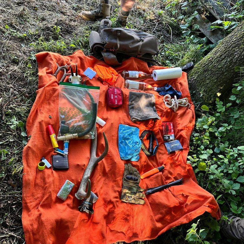 Verschiedene Outdoor-Ausrüstung auf einer orangefarbenen Decke, einschließlich Erste-Hilfe-Kit, Werkzeuge, eine Flasche und ein Schädel. Ein Rucksack und Schuhe sind in der Nähe.