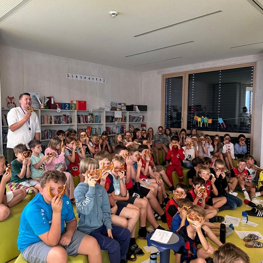 Gruppe von Kindern isst Brezeln im Klassenzimmer. Ein Lehrer hält eine Brezel vor den Schülern. Bücherregale und eine Tafel sind im Hintergrund.