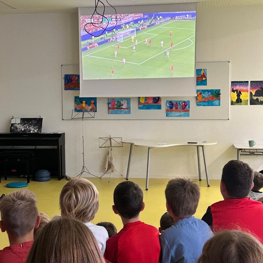 Mehrere Kinder in einem Klassenzimmer sehen sich ein Fußballspiel auf einer großen Projektionsleinwand an der Wand an. Ein Klavier, ein Tisch und verschiedene Kunstwerke schmücken den Raum.