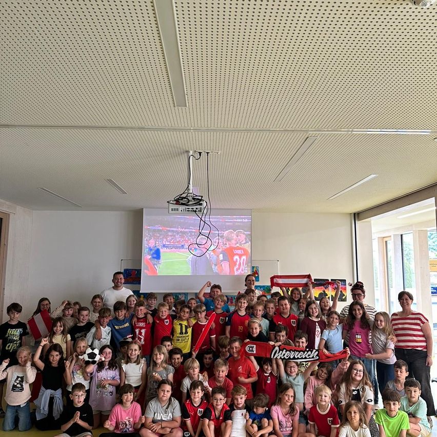 Eine Gruppe von Kindern und Erwachsenen versammelt sich in einem Raum mit einer Projektionsleinwand und schaut sich ein Fußballspiel an. Einige halten eine rote Fahne und einen Fußball.