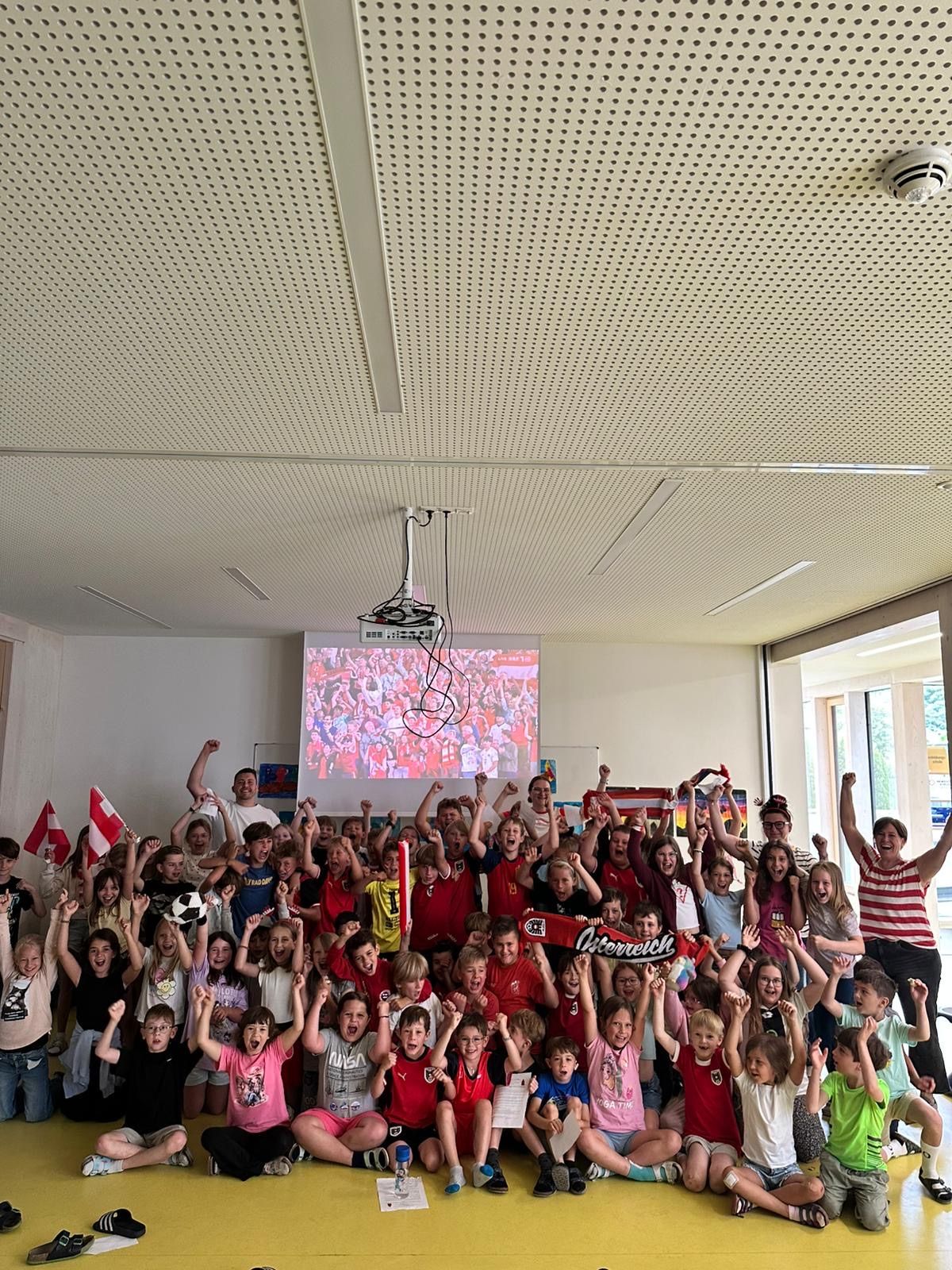Eine Gruppe von Kindern in einem Klassenzimmer, viele in roten Shirts, jubeln mit Flaggen und Bannern und schauen sich eine Sportveranstaltung auf einer Projektionsleinwand an.