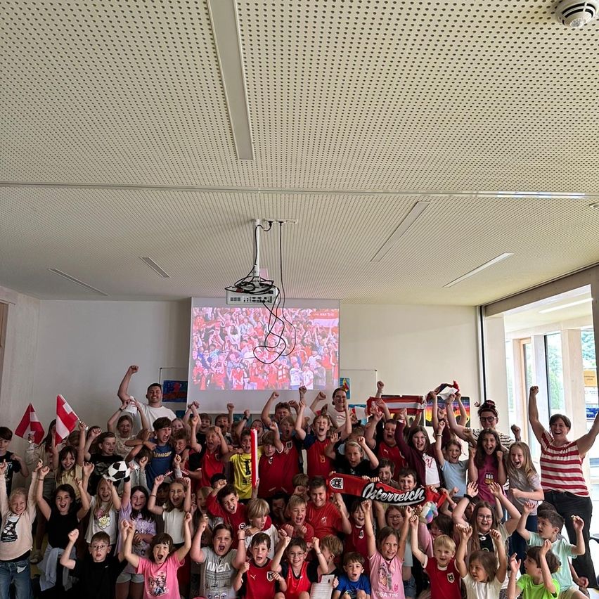 Eine Gruppe von Kindern in einem Klassenzimmer, viele in roten Shirts, jubeln mit Flaggen und Bannern und schauen sich eine Sportveranstaltung auf einer Projektionsleinwand an.