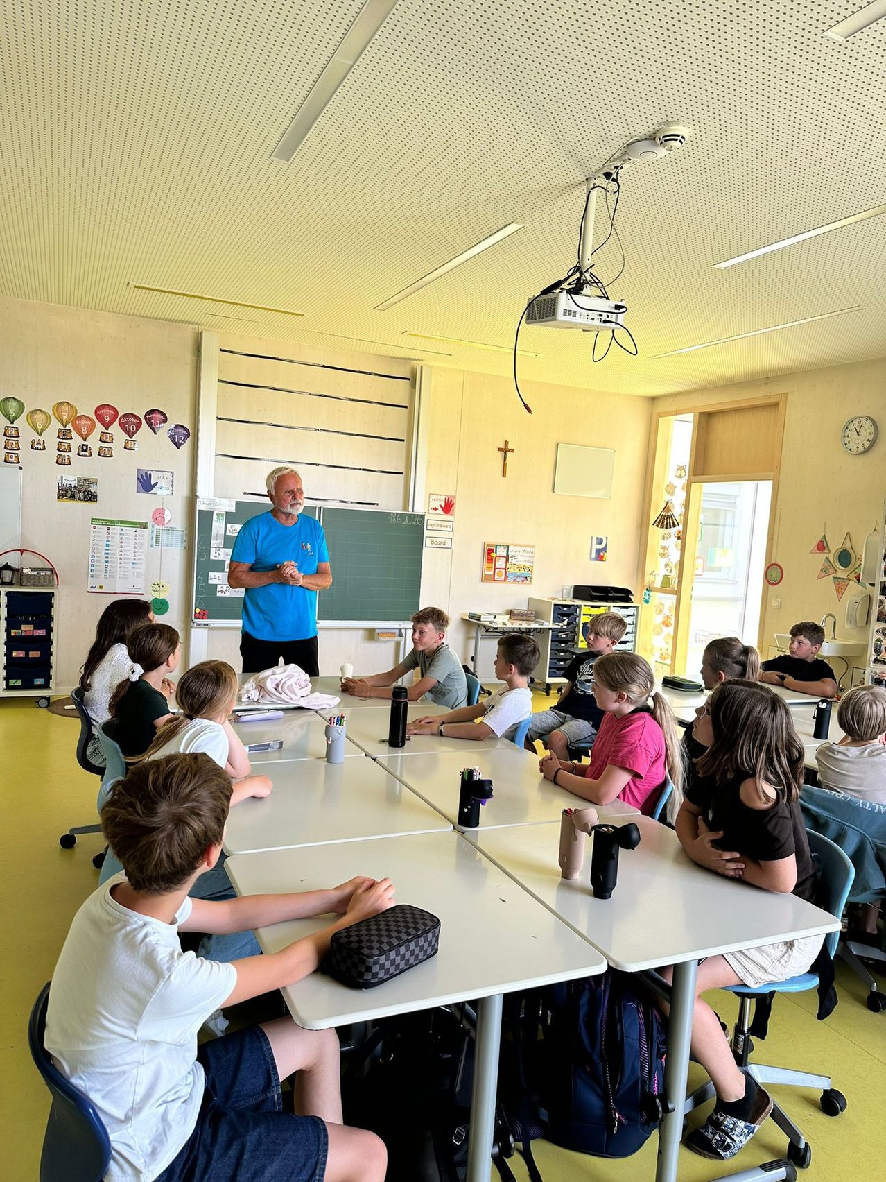 Ein Klassenzimmer mit Schülern, die an Tischen sitzen, ein Mann in einem blauen Shirt steht vor einer Tafel. Verschiedene Gegenstände wie ein Projektor und eine Uhr sind vorhanden.