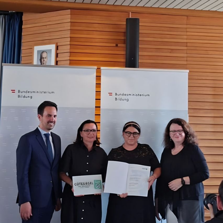 Ein Mann und drei Frauen stehen vor einer Schriftrolle, die 'Bundesministerium Bildung' lautet, halten Urkunden und Dokumente in den Händen. Der Mann ist schick gekleidet in Anzug und Krawatte.