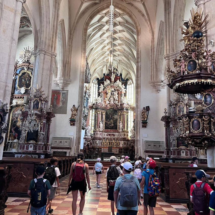 Eine Gruppe von Touristen besucht eine prunkvolle gotische Kirche mit hohen Bögen und aufwendigen Verzierungen. Einige tragen Hüte und haben Rucksäcke dabei.