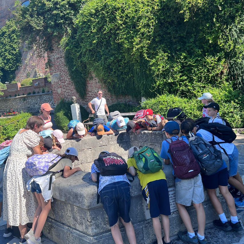 Eine Gruppe von Kindern und Erwachsenen lehnt sich über eine Steinmauer, trägt Hüte und Rucksäcke. Dahinter steht ein Mann neben einer Statue und einer Mauer.