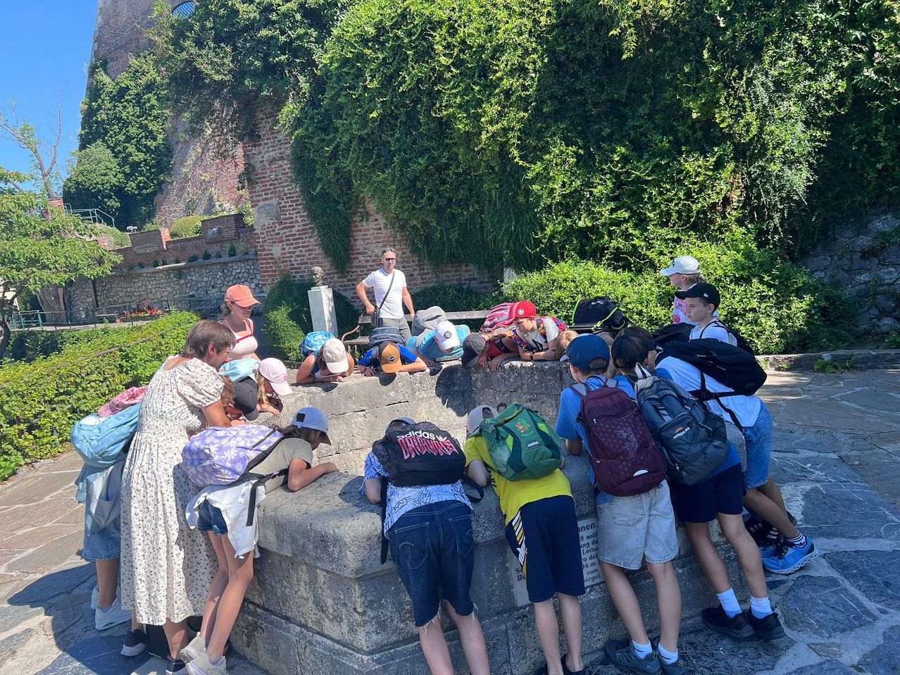 Eine Gruppe von Kindern und Erwachsenen lehnt sich über eine Steinmauer, trägt Hüte und Rucksäcke. Dahinter steht ein Mann neben einer Statue und einer Mauer.