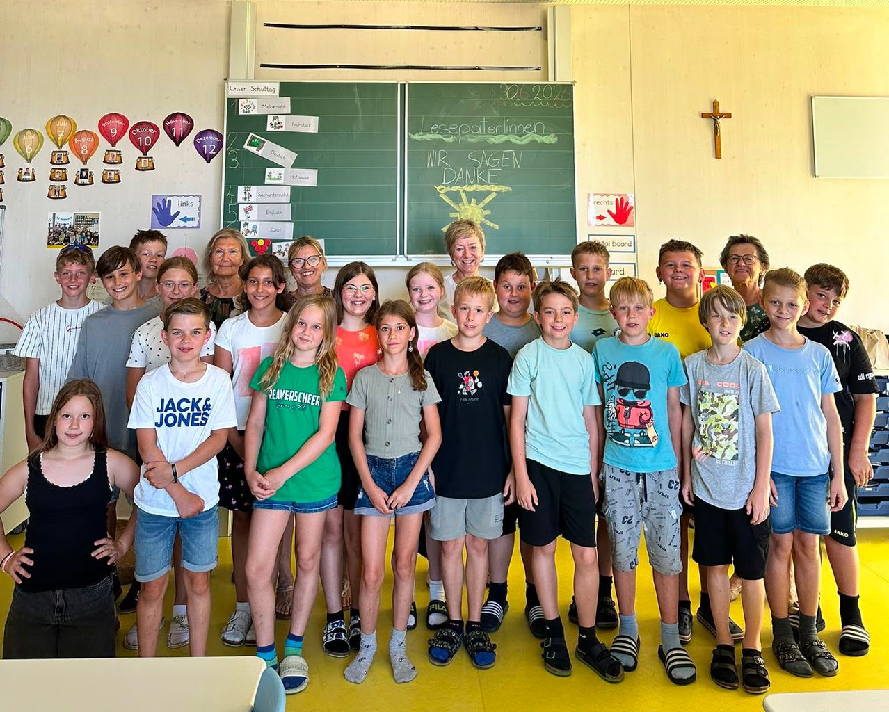 Eine Gruppe von Schülern und Lehrern steht vor einer Tafel mit einer gelben Sonne und den Worten 'Wir sagen Danke.' Der Raum hat bunte Dekorationen und ein Kreuz an der Wand.
