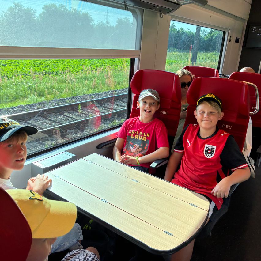 Vier Kinder sitzen in einem Zugabteil und lächeln. Ein Junge trägt ein rotes Fußballtrikot, ein anderer einen Hut. Das Fenster zeigt ein Grasfeld und Gleise.
