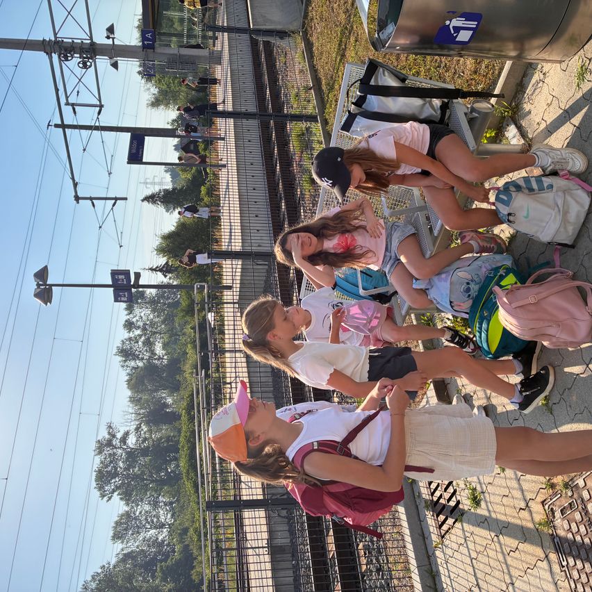 Eine Gruppe von Mädchen mit Rucksäcken wartet auf einem Bahnsteig. Sie sitzen auf dem Boden und lehnen sich gegen einen Zaun. Bäume und Stromleitungen sind im Hintergrund.